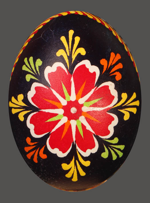 Jajko-pisanka, pośrodku czerwony kwiat z białymi brzegami płatków, wewnątrz żółto-biało-pomarańczowa rozeta. po bokach zielone, żółte, czerwone jodełki, tło czarne