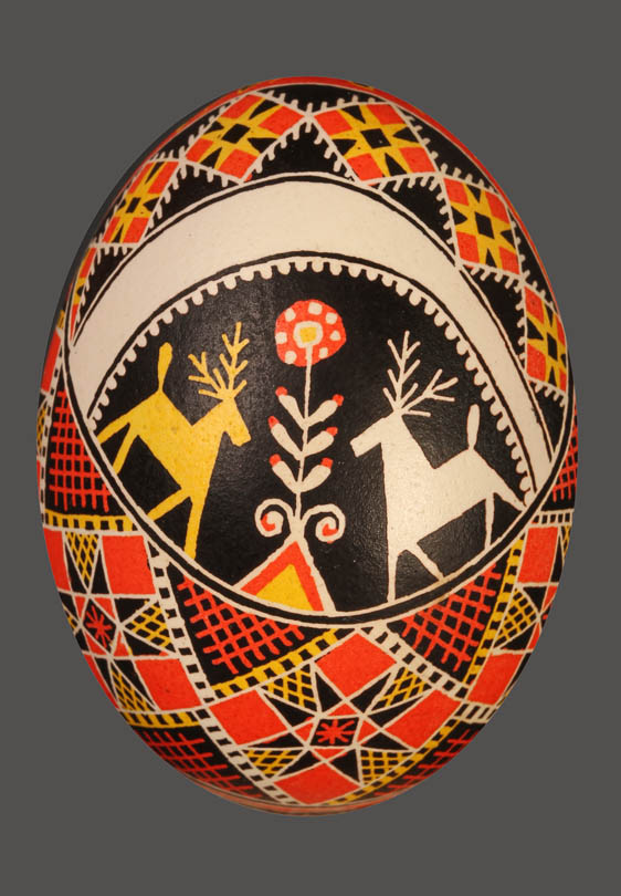 Jajko-pisanka z drzewkiem zycia pośrodku i dwoma jelonkami po bokach (bialy i żółty), poniżej i powyżej ornamenty geometryczne (czerwony, zółty, biały i czarny kolor); tło czarne