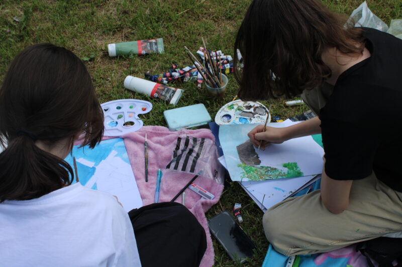 Dwie dziewczynki siedzą na trawie. Malują drewniane chałupy,