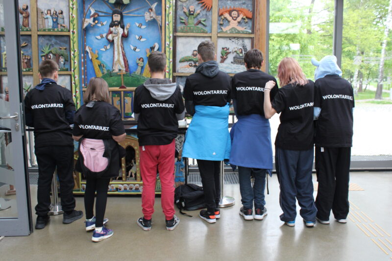 Grupa wolontariuszy stoi tyłem do fotografa. Dzieci prezentują koszulki z napisem 