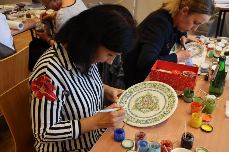 Uczestniczka-konkursu-siedzi-przy-stole-i-maluje-duzy-plaski-talerz-w-ornamenty-roslinno-kwiatowe.
